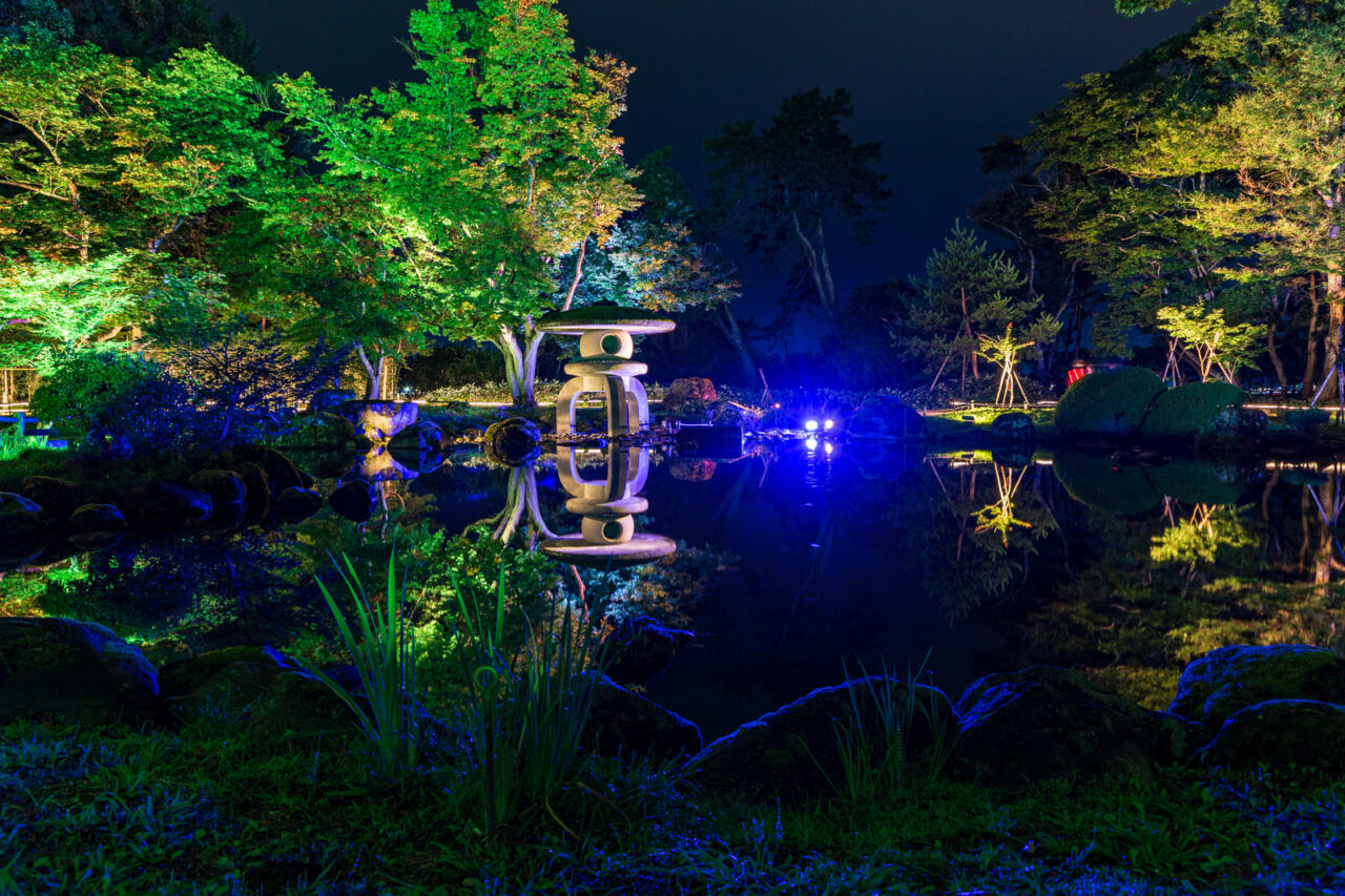 照明に照らされた木々と石灯籠が夜の池に映る日本庭園